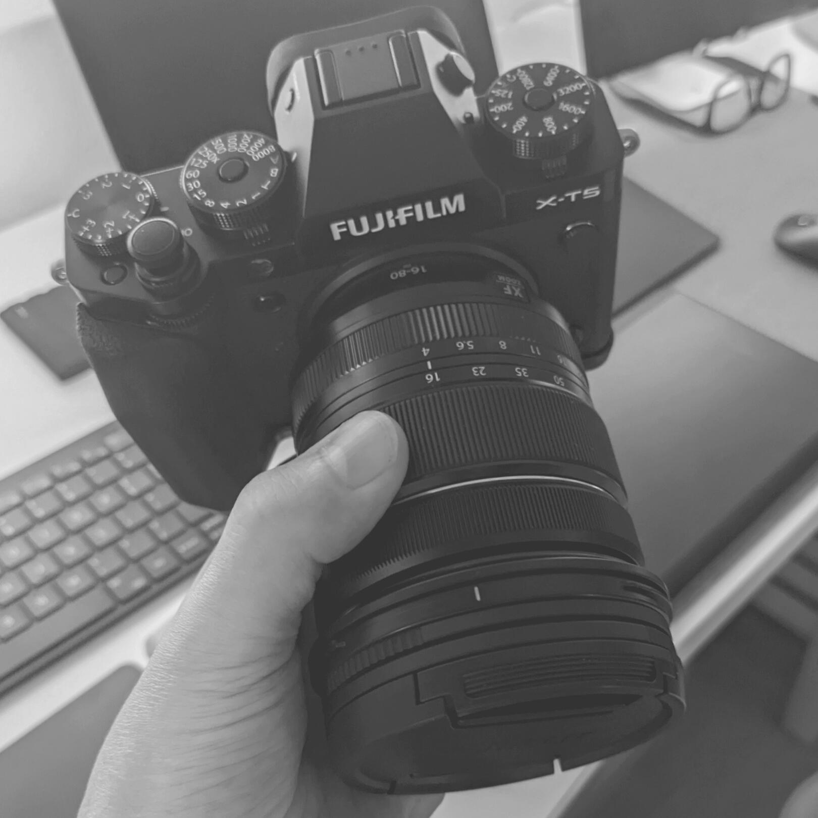 A monochrome snapshot of a hand holding a black Fujifilm X-T5 camera with a Fujinon 16-80mm lens.