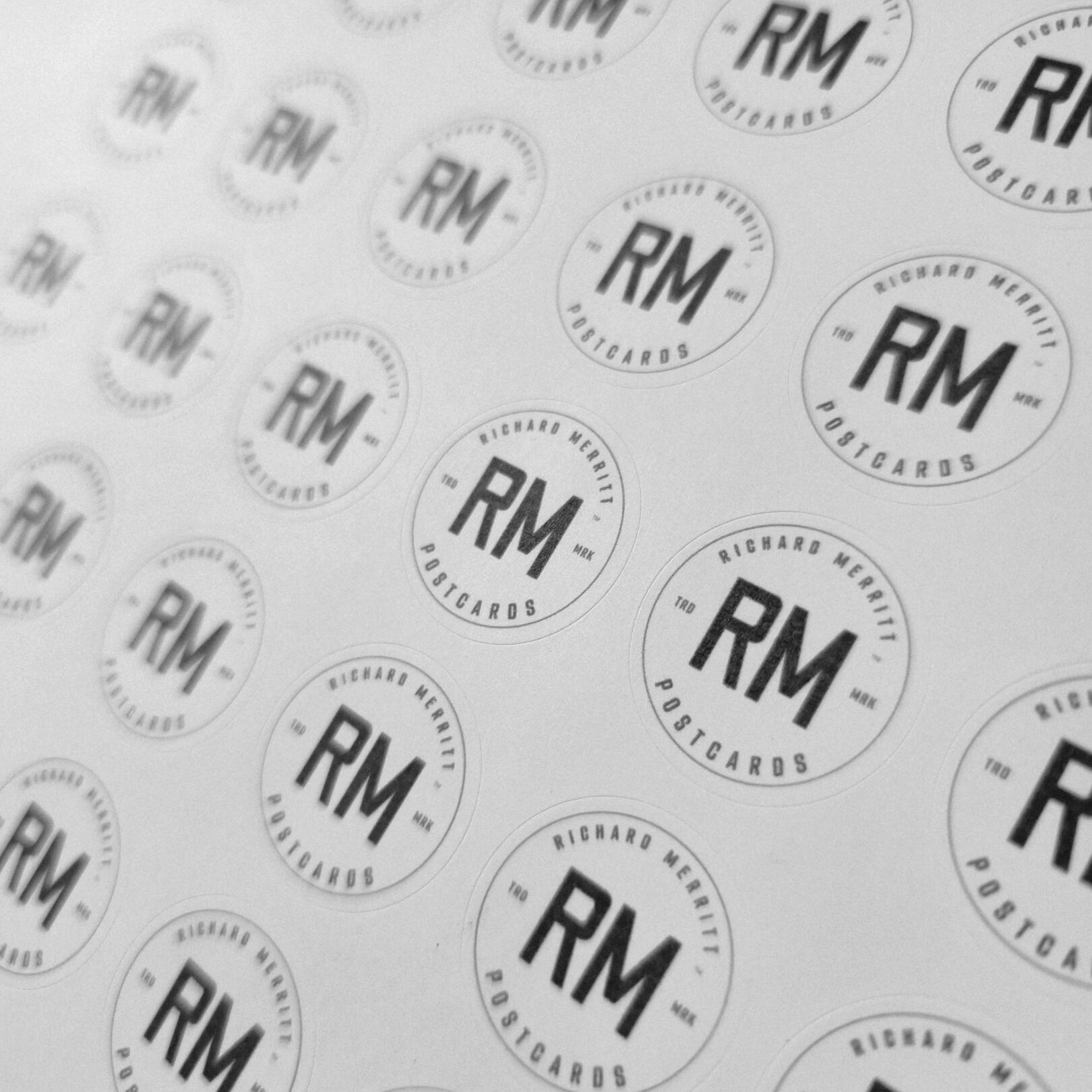 A close-up monochrome snapshot of a circular logomark badge in black ink repeated on a sheet of sticker paper. The logomark reads: "RM - Richard Merritt Postcards".