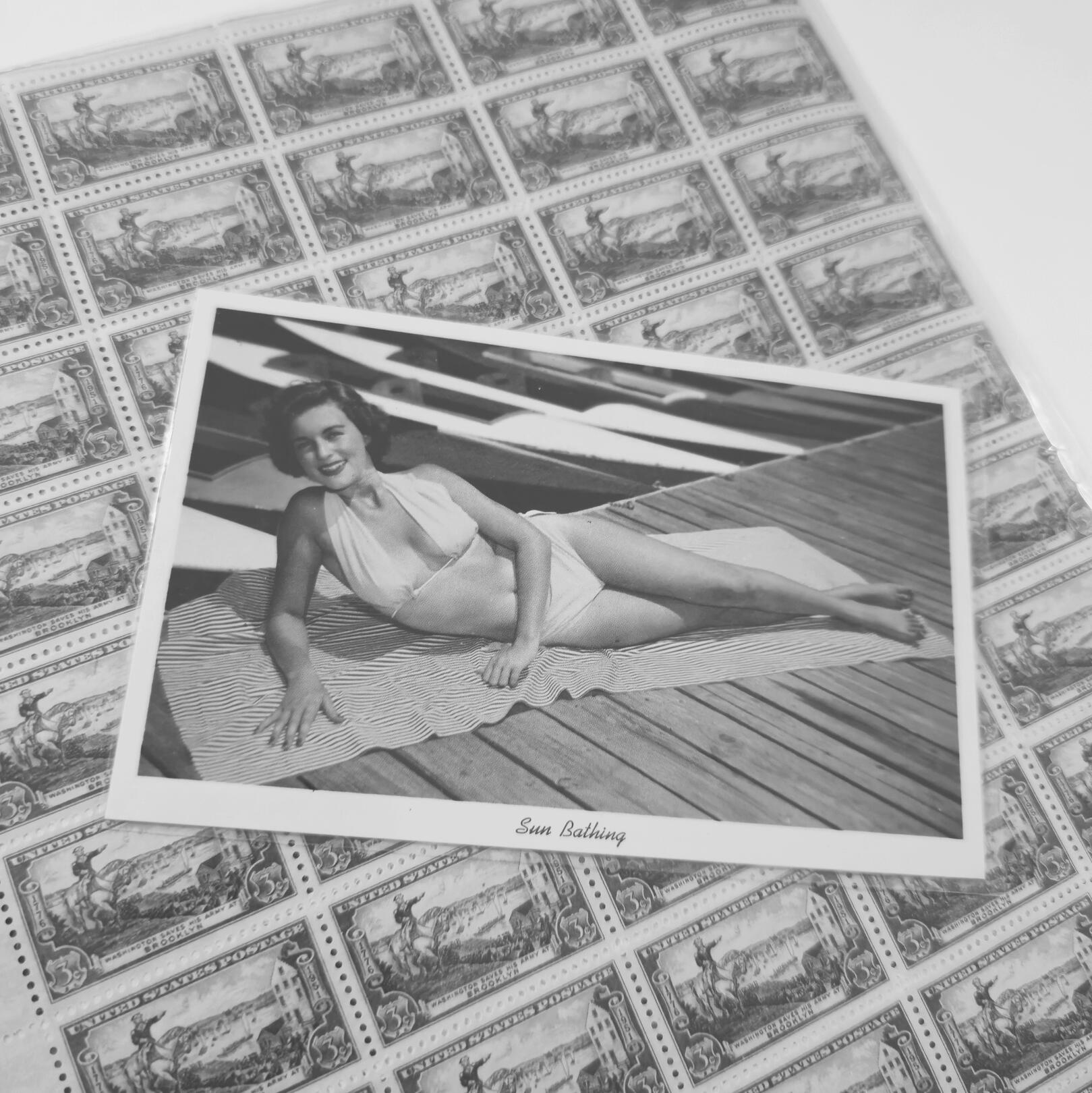 A close-up monochrome snapshot of a vintage circa 1953 Curt Teich Co. pin-up postcard of a woman in a bikini sunbathing on a boat dock. The postcard is sitting on a USPS postage stamp full-sheet: 1951 Battle of Brooklyn 3¢ stamp archived for RM Postcards b