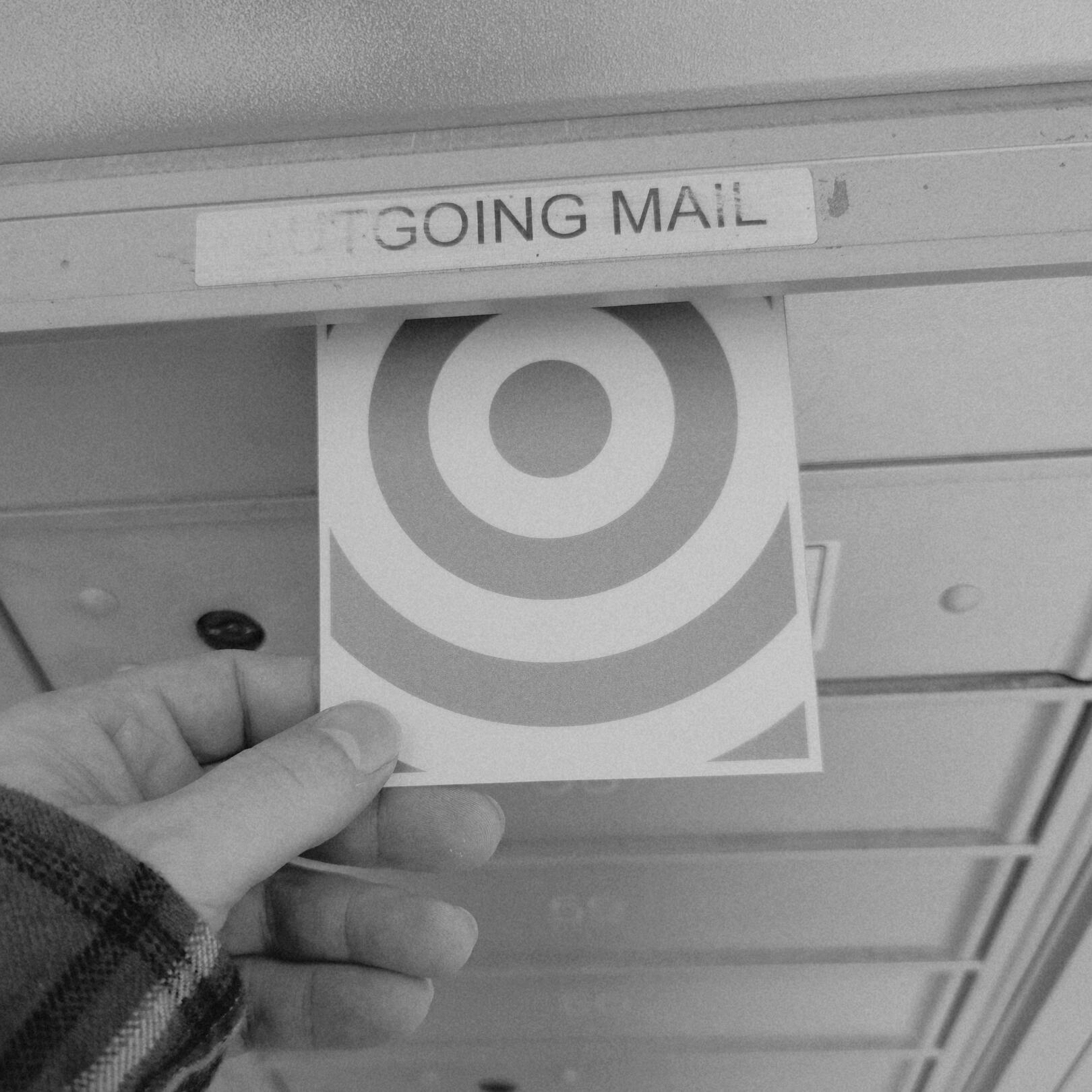 A close-up monochrome snapshot of a Richard Merritt | RCHRDMRRTT Limited Edition postcard (with a bullseye on the front) being dropped into the outgoing mail slot of a postal box for a Postcrossing mailing to Taiwan.