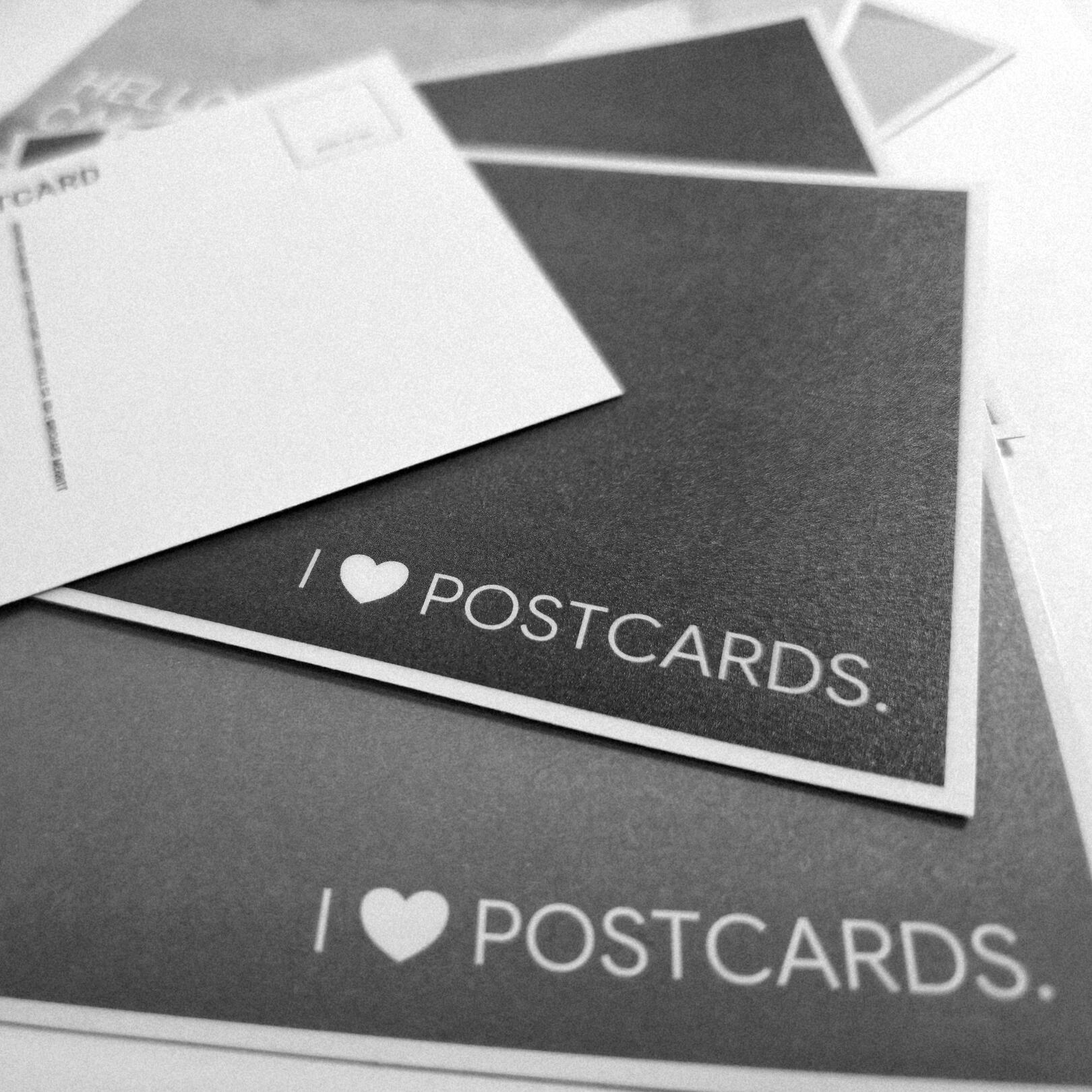 A close-up monochrome snapshot of several Limited Edition Postcards by Richard Merritt. Two of the postcards read: I ♥ POSTCARDS.