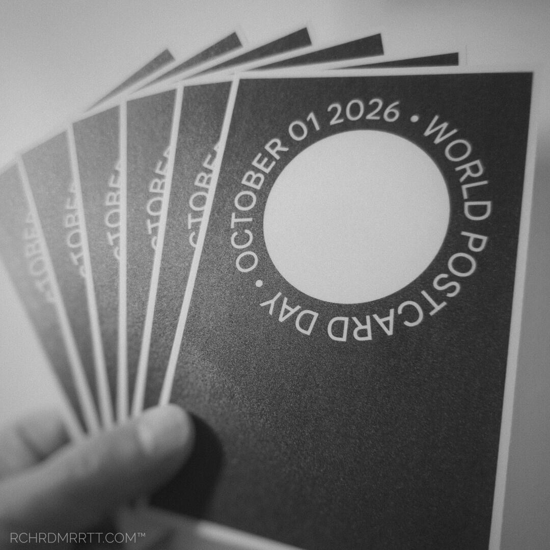 A monochrome square snapshot of a hand holding a fanned-out stack of World Postcard Day postcards designed and printed by Richard Merritt from California USA.